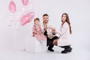 Genç ebeveynler, anne ve baba iki yaşındaki çocuklarının doğum gününü kutluyorlar. Aile ışığı ve pastadaki mumları söndürmek. Arka planda pembe toplarla süslenmiş çiçekler..
