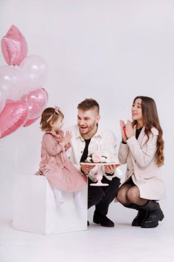 Genç ebeveynler, anne ve baba iki yaşındaki çocuklarının doğum gününü kutluyorlar. Aile ışığı ve pastadaki mumları söndürmek. Arka planda pembe toplarla süslenmiş çiçekler..