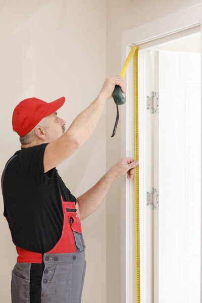 Worker in red cap and work suit with the measuring tape. Man is installing the doors. Measure tape in hands. Repair works. Maintenance in the apartment