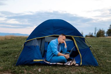 Adam doğadaki çadırda dizüstü bilgisayarda çalışıyor. Kampta oturan genç bir serbest yazar. Dağlarda dinleniyorlar. Uzaktan çalışma, yazın açık hava aktivitesi. Mutlu erkek işi tatilde.