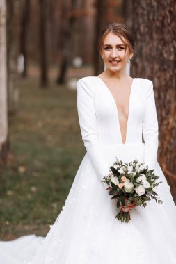 Uzun beyaz elbiseli gülümseyen gelin parkta duruyor ve elinde gül çiçekleri ve yeşilliklerden oluşan bir buket tutuyor. Saç stili, makyaj ve otel yakınlarındaki açık havada uzun peçeli genç bir kadın..