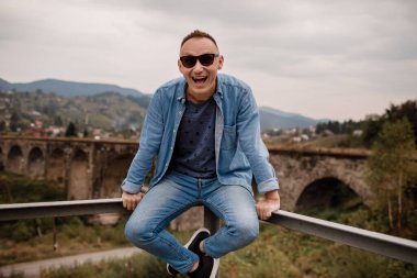 Vorokhta, Karpatlar 'daki demiryolu köprüsündeki şık genç adam bir dağ manzarasının arka planına karşı. Demiryolu yakınlarındaki dağlarda oturan turistler..