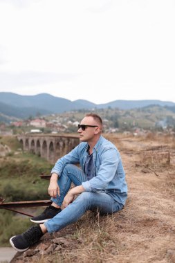 Vorokhta, Karpatlar 'daki demiryolu köprüsündeki şık genç adam bir dağ manzarasının arka planına karşı. Demiryolu yakınlarındaki dağlarda oturan turistler..