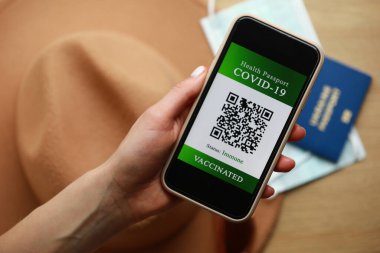 Yeni normal konsept. Kadının elindeki görüntüyü kapat. Bağışıklık sistemi dijital pasaportlu bir akıllı telefon. Arka planda, güneş gözlüğü, koruyucu maske, şapka ve pasaport. bir bulanıklık içinde