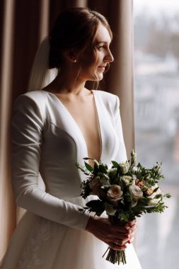 Gelin, otel odasının penceresinin yanında bir buket çiçekli beyaz gelinlik içinde. Saç stili ve makyajı olan mutlu, güzel bir kadın. düğün günü.