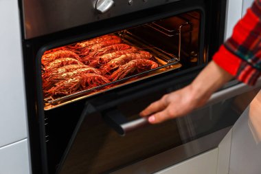 Ev mutfağında ızgara karides ve kızarmış karides. Kadın paslanmaz tepside karides barbeküsü yapıyor ve gece partisini kutlamak için elektrikli fırında yemek pişiriyor. deniz ürünleri.