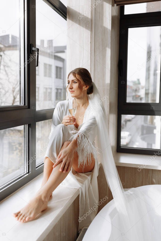 Bride s morning. Bride drinking champagne in the peignoir. young woman is sitting on a large window in a hotel room in bathroom. Beautiful girl in white wedding robe. wedding day.