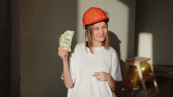 Gelukkige jonge glimlachende vrouwelijke werknemer in oranje hardhat met geld, staande op de bouwplaats in appartement. Architect houdt geld bij voor het huisproces.. — Stockvideo