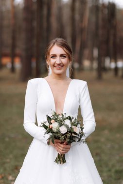 Uzun beyaz elbiseli gülümseyen gelin parkta duruyor ve elinde gül çiçekleri ve yeşilliklerden oluşan bir buket tutuyor. Saç stili, makyaj ve otel yakınlarındaki açık havada uzun peçeli genç bir kadın..