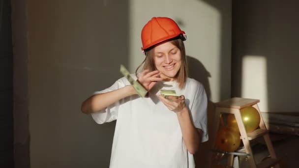 Молода усміхнена жінка-працівник в помаранчевому жорсткому капелюсі рахує гроші, стоячи на будівельному майданчику в місті. Архітектор кидає і грає з доларами в офісі . — стокове відео