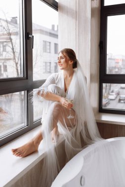 Gelinin sabahı. Gelin, Peignoir 'de şampanya içiyor. Genç bir kadın banyodaki bir otel odasında büyük bir pencerede oturuyor. Beyaz gelinlikli güzel kız. düğün günü.