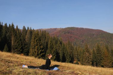 Güneşli dağların arka planında yoga minderi üzerinde egzersiz yapan formda genç bir kadın. Açık hava sporu, sağlıklı yaşam tarzı. Spor kıyafetli bir kadın sabahları ormana karşı doğada asana yapıyor..