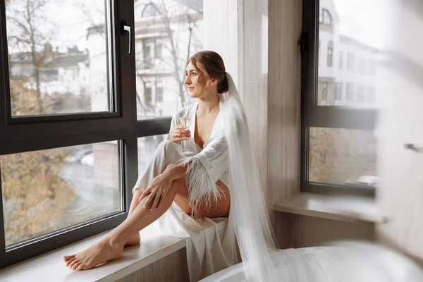 花嫁の朝 花嫁はペノワールでシャンパンを飲む 若い女性が浴室のホテルの部屋の大きな窓の上に座っている 白いウェディングローブの美しい女の子 結婚式の — ストック写真