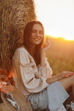 Genç kadın yaz günbatımının güzel ışığında bir tarlada saman balyalarının yanında oturuyor. Sahada uzun saçlı, güzel romantik bir kız..