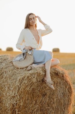 Uzun saçlı, kot etek giyen, elinde açık tişört ve saman torbasıyla yaz aylarında balyada oturan genç bir kadın. Doğal kırsal kesimde bir kadın portresi. Çevre eko turizmi kavramı