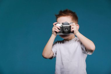 Küçük çocuk retro kamera kullanarak fotoğraf çekiyor. Klasik fotoğraf makinesi olan çocuk mavi arka planda izole edilmiş. Kopya uzayı ile eski teknoloji kavramı. Çocuk fotoğrafçılığı öğreniyor .