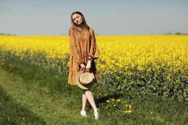 Güzel genç bir kadın sarı kolza tohumu tarlasında yürüyor. Esmer, uzun saçlı, rüzgarda uçuşan bir kız. Elbise ve hasır şapka giymiş. Yaz tatili konsepti.