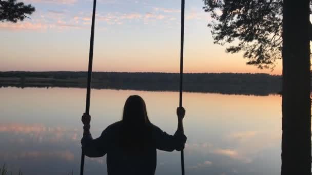 夕暮れの湖の上で揺れるロマンチックな若い女性のシルエット。美しい自然の中でスイングに座っている若い女の子の旅行者,湖の上に表示 — ストック動画