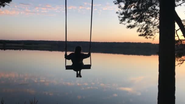 夕暮れの湖の上で揺れるロマンチックな若い女性のシルエット。美しい自然の中でスイングに座っている若い女の子の旅行者,湖の上に表示 — ストック動画
