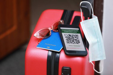 Yeni normal konsept. Bağışıklık sistemi olan dijital bir sağlık pasaportu, kırmızı bavulu, koruyucu maskesi, pasaportu ve güneş gözlüğü olan bir akıllı telefonun yakın görüntüsü. Bir salgın sırasında uçakla seyahat etmek