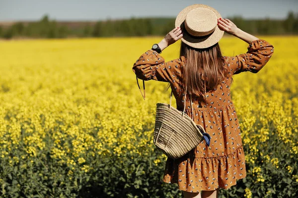 Rear view. beautiful young woman in a dress holding a hat and walking in a rapeseed field for summer, view from the back. copy space. summer holiday concept.