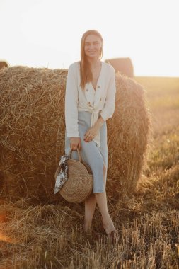 Kot etekli ve elinde saman torbasıyla gülümseyen güzel bir esmerin tam boy portresi. Güneşli yaz gününde saman balyalarıyla buğday tarlasında yürüyüş yapan bir kadın.