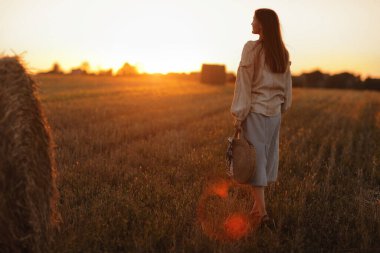 Genç bir kadın yaz günbatımının güzel ışığında bir tarlada saman balyalarının yanında yürüyor. Sahada uzun saçlı, güzel romantik bir kız.