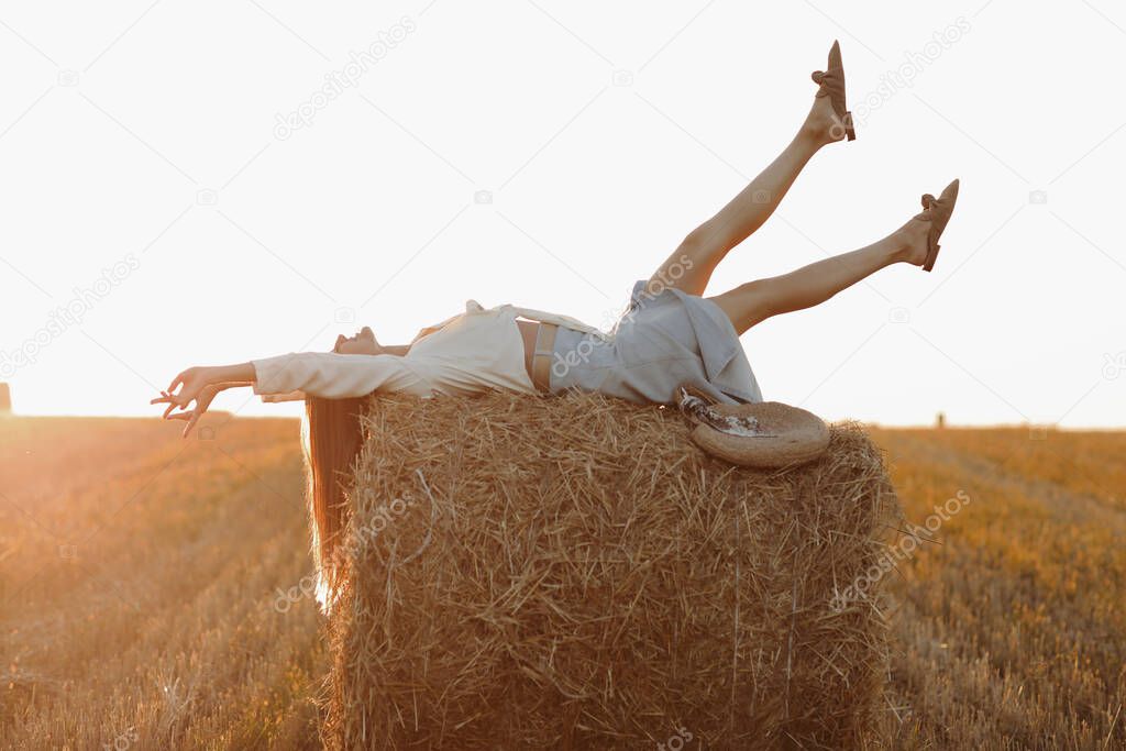 Young woman with long hair, wearing jeans skirt, light shirt is lying on straw bale in field in summer on sunset. Female portrait in natural rural scene. Environmental eco tourism concept