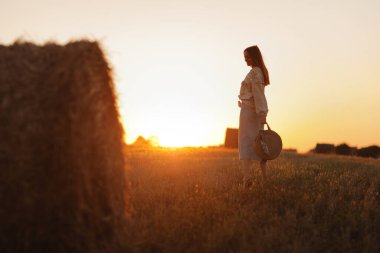 Genç bir kadın yaz günbatımının güzel ışığında bir tarlada saman balyalarının yanında yürüyor. Sahada uzun saçlı, güzel romantik bir kız.
