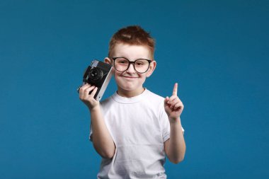 Küçük çocuk retro kamera kullanarak fotoğraf çekiyor ve parmağını kaldırıyor. Klasik fotoğraf makinesi olan çocuk mavi arka planda izole edilmiş. Eski teknoloji konsepti. Çocuk fotoğrafçılığı öğreniyor.