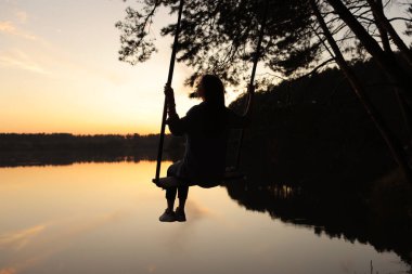 Gün batımında gölde sallanan romantik genç bir kadının silüeti. Genç bir gezgin kız salıncakta oturuyor, güzel bir tabiatı var, gölü görüyor..