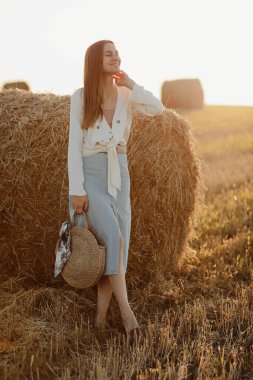 Kot etekli ve elinde saman torbasıyla gülümseyen güzel bir esmerin tam boy portresi. Güneşli yaz gününde saman balyalarıyla buğday tarlasında yürüyüş yapan bir kadın.