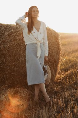 Kot etekli ve elinde saman torbasıyla gülümseyen güzel bir esmerin tam boy portresi. Güneşli yaz gününde saman balyalarıyla buğday tarlasında yürüyüş yapan bir kadın.