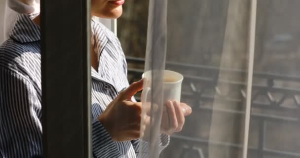 4 k. jovem bonita feliz com uma toalha na cabeça está tomando café da manhã na varanda, bebendo café ou chá. menina olha para fora sobre os telhados da cidade hora da manhã — Vídeo de Stock