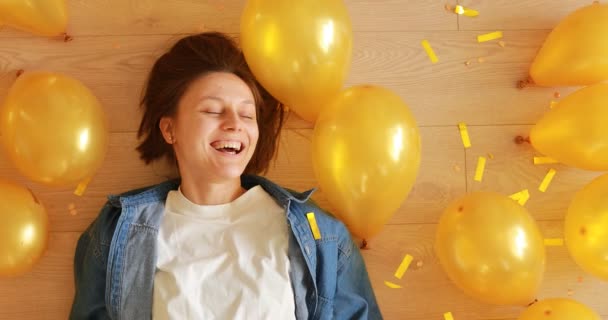 Szczęśliwa młoda kobieta na przyjęciu urodzinowym uśmiechnięta i bawiąca się leżeniem na podłodze ze złotym konfetti i balonami — Wideo stockowe