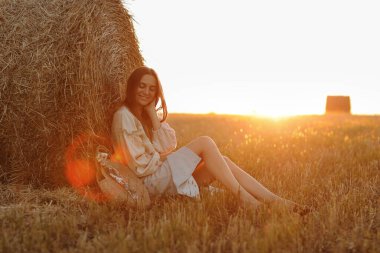 Genç kadın yaz günbatımının güzel ışığında bir tarlada saman balyalarının yanında oturuyor. Sahada uzun saçlı, güzel romantik bir kız..