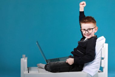 Gözlüklü okul çocuğu dizüstü bilgisayar kullanarak yatakta oturuyor. Mutlu çocuk okul ödevini yapıyor ve sevinçle ellerini kaldırıyor. Çevrimiçi öğrenim, uzaklık dersi, ev izolasyonunda online öğretmenle eğitim.