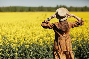Dikiz aynası. Şapkalı, kolza tohumu tarlasında yaz boyunca yürümüş, arkadan manzaralı güzel bir genç kadın. kopyalama alanı. Yaz tatili konsepti.