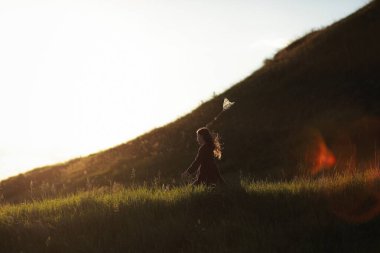 Küçük kız kelebek ağıyla yürüyor ve güneşli yaz gününde yeşil tepelerde kelebekler yakalıyor. Boşluğu kopyala.
