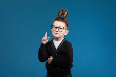 Mutlu Paskalyalar çocuklar. Tavşan kulaklı çocuk mavi stüdyo arka planında gözlüklü. İşaret ediyor. Kameraya bakıp gülümsüyor.