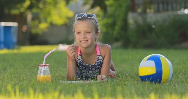 Девочка-подросток в купальнике и солнечных очках ест картошку фри и пьет апельсиновый сок на зеленой лужайке во время летнего пикника. на заднем плане бассейна. Летние каникулы. — стоковое видео