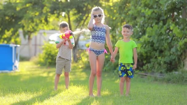 Tre roliga barn pojkar och flicka leker med vattenpistoler på grön gräsmatta i trädgården på varm sommardag i slow motion. simbassäng på bakgrund. — Stockvideo