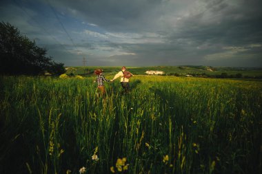 Sevgi dolu hipster bir çift sahada yürüyor, öpüşüyor ve el ele tutuşuyor, sarılıyor, yazın günbatımında çimlerin üzerinde uzanıyor. Sevgililer Günü.