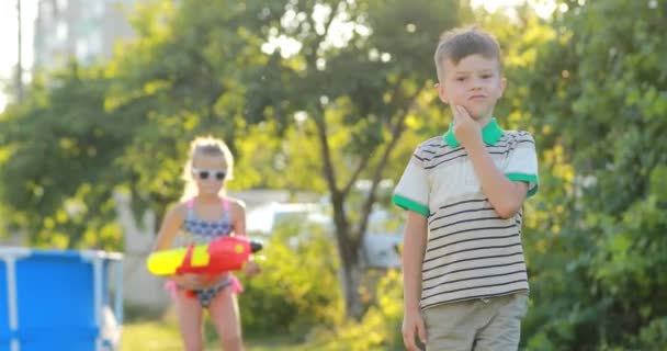 Två roliga barn pojke och flicka leker med vattenpistoler på grön gräsmatta i trädgården på varm sommardag i slow motion. simbassäng på bakgrund — Stockvideo