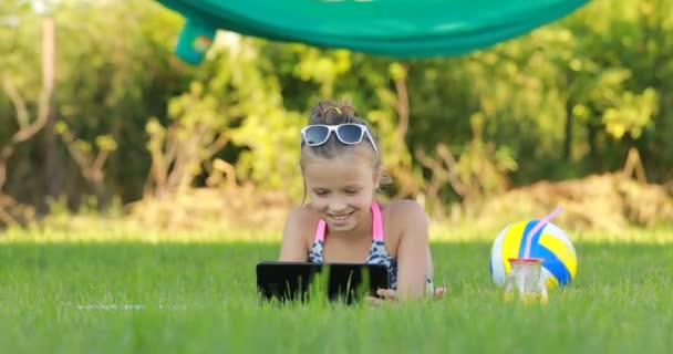 Dívka v sunglasess používá notebook a leží na trávě v letní slunečný den. Houpací síť a míč na pozadí. letní dovolená. — Stock video