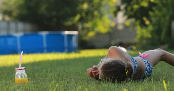 Adolescente chica en un traje de baño y gafas de sol miente y haning diversión en el césped verde, mientras que las vacaciones de verano en el fondo de la piscina. jugo con una pajita en un frasco. Actividad de verano — Vídeo de stock