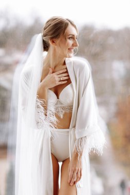 Düğünden önce gelinin sabahı. Uzun peçeli, beyaz cüppeli ve pencerede geceliği olan güzel bir kadın. Doğal güzellik ve profesyonel makyaj. Doğal ışık