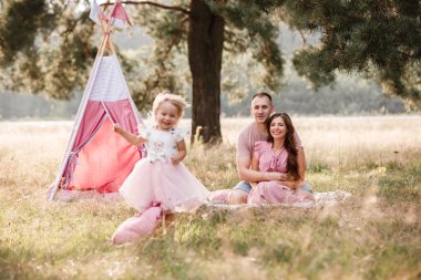 Anne, baba ve küçük kız parkta wigwam dekorasyonunun yanında oturuyorlar. Aile yazın dışarıda vakit geçirir, birlikte eğlenir. Pembe elbiseli kız..