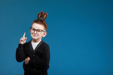 Mutlu Paskalyalar çocuklar. Tavşan kulaklı çocuk mavi stüdyo arka planında gözlüklü. İşaret ediyor. Kameraya bakıp gülümsüyor..