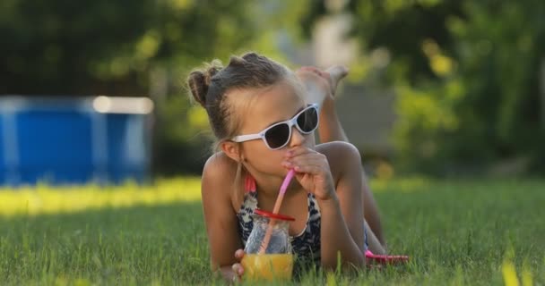 Маленькая девочка в купальнике и солнцезащитных очках пьет коктейль с соломинкой. Счастливый, милый ребенок лежит на траве. Делай лица и улыбайся. Летние каникулы. — стоковое видео